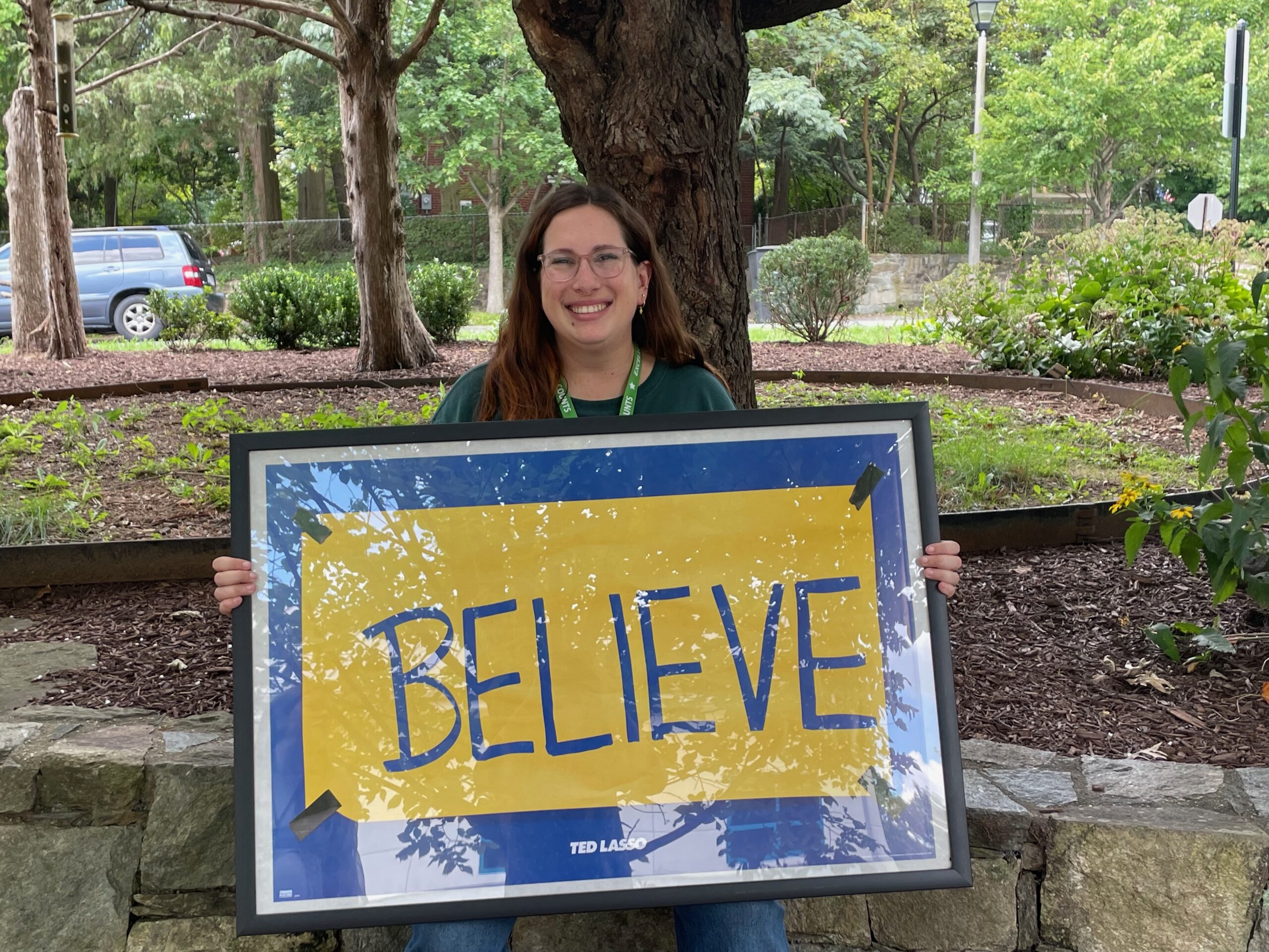 Photo of Ms. Mintzer holding a sign that says "BELIEVE" from the tv show Ted Lasso.